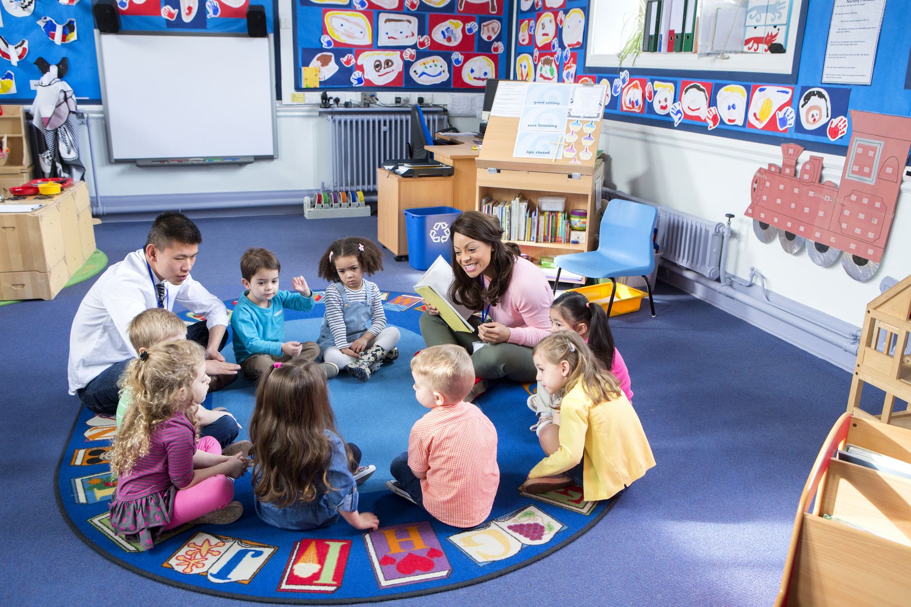 Group of nursery children sitting on the floor in their classroom with their teachers. The female teacher is reading from a book.